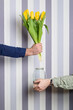 A man's hand with a bouquet of yellow tulips and a woman's hand with a jar of water on a striped background.