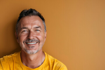 Wall Mural - A man with a beard and gray hair is smiling at the camera. He is wearing a yellow shirt and he is happy. Happy smiling middle aged adult man on a solid yellow background