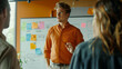 A man in an orange shirt is gesturing during a team meeting with a whiteboard in the background.
