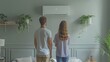 Standing in the living room, a young couple is gazing at the white air conditioner against the grey wall.