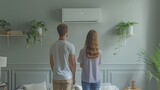 Fototapeta Na sufit - Standing in the living room, a young couple is gazing at the white air conditioner against the grey wall.