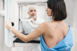 © illustrissima - man doctor stands against the background of patient who is doing a mammogram. early diagnosis of breast cancer. mammography machine. routine regular examination for women over 35 years of age