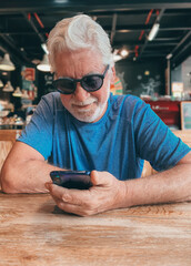 Wall Mural - Handsome smiling bearded senior man sitting at cafe table using mobile phone browsing internet