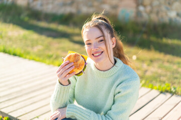 Poster - Young pretty girl holding a burger at outdoors smiling a lot