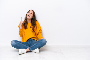 Canvas Print - Young caucasian woman sitting on the floor isolated on white wall pointing up and surprised