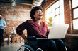 African American businesswoman in a wheelchair laughs while working on her laptop in a creative office space, embodying success and accessibility.

