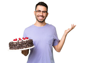 Young caucasian man holding birthday cake isolated on green chrome background extending hands to the side for inviting to come