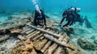 Two divers in scuba gear closely inspect a wooden plank resting on the ocean floor