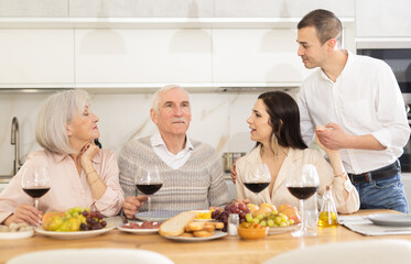 Senior couple gathered at kitchen table with grown son and daughter-in-law, enjoying conversation with wine and light snacks..