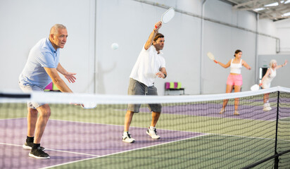 Wall Mural - Portrait of sporty senior man playing doubles pickleball with male partner on indoor court, ready to hit ball. Sport and active lifestyle concept..