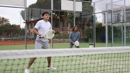 Wall Mural - Portrait of emotional determined young guy playing padel tennis on open court in summer, swinging racket to return ball over net. Sportsman ready to hit volley