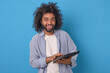 Young cheerful curly Arabian business man with tablet computer in hands looks at screen with smile inviting you to use gadgets for distance learning or work stands on blue studio background.