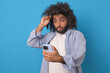 Young shocked optimistic Arabian man lifts glasses from eyes in surprise reading fake news on phone and misinformation sent to him via internet stands on plain blue background. Mobile, smartphone