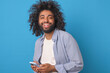 Young carefree Arabian man student laughing holding mobile phone and using instant messengers to chat with girlfriend or exchange funny pictures with college buddies stands on blue background.