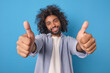 Young smiling attractive Arabian man shows thumbs up stretching arms towards camera as sign of approval of political program of party before elections dressed in casual shirt stands in blue studio.