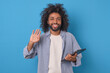 Young positive ethnic Arabian man entrepreneur waves hand friendly and holds tablet computer to do online work on freelance exchange dressed in casual clothes stands on plain blue background.