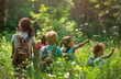 A teacher and group of children were on an outdoor field trip in nature, learning about plants from the female naturalist smiling at the camera, standing among tall grasses with backpacks beside them