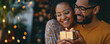 A couple is holding a gold wrapped gift, smiling at the camera. Scene is happy and festive, as it is a Christmas or holiday gift