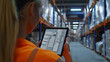 A female warehouse worker wearing an orange shirt is focused on looking at a tablet device.