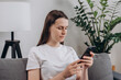 Happy cute young woman sitting on cozy couch using cell phone, smiling lady laughing holding smartphone, looking at cellphone enjoying doing online ecommerce shopping in mobile apps or watching videos