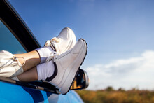A Close-up Of A Female Legs In White Sneakers Are Exposed To The Car Window. Traveling By Automobile. Blue Sky And A Field In The Background.