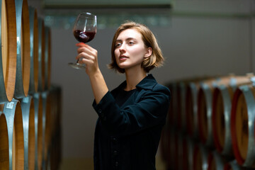 Professional Caucasian woman sommelier tasting and sniffing red wine in wine glass at wine cellar with wooden barrel. Winery brewery liquor wine factory manufacturing industry and winemaker concept. 