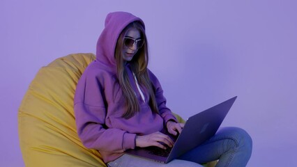 Young woman sitting on pouf and using notebook isolated on violet color background. Girl in hoodie working remote and lying on beanbag. Senior level program developer wearing sunglass during daily