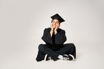 Wall Mural - smiling charming guy in graduate outfit sitting and holding with hands to cheeks on grey background