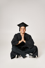 Wall Mural - attractive young guy in graduate gown and cap sitting and folding hands in grey background