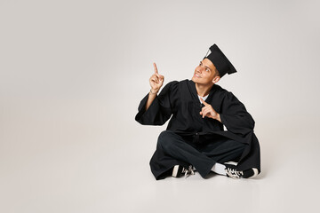 Wall Mural - cheerful young man in graduate gown and cap sitting and showing with fingers to up