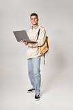 Fototapeta Młodzieżowe - young student in headphones standing with backpack and networking to laptop against grey background