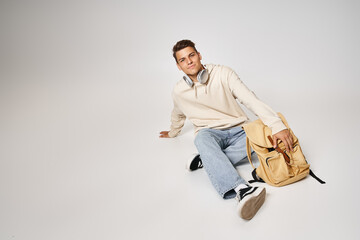 Wall Mural - handsome young man in headphones and casual outfit sitting with backpack on grey background