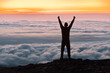 Hiker silhouette is standing in winner pose at mountain top and looks at sunrise above the clouds
