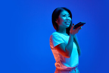 Beautiful Asian woman using phone and looking away against blue gradient background in neon light. Cellular connection. Concept of business, study and work, startup, sales, shopping.