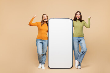 Wall Mural - Full body young friend two women they wear orange green shirt casual clothes together big huge blank screen mobile cell phone smartphone do winner gesture with area isolated on plain beige background