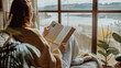 © Kanlayarawit - A woman wearing glasses is reading a book while sitting on a couch with a view of a lake. Concept of relaxation and tranquility, as the woman enjoys her book in a peaceful setting. Winter season.