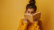 A sad white teenage girl is reading a white book on a plain yellow background with copy-space for text.