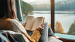 A woman is reading a book while sitting on a couch with a view of a lake. Concept of relaxation and tranquility, as the woman enjoys her book in a peaceful setting. Evening time with mountain view.