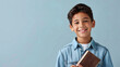 A happy and smiling Latino boy is holding a brown book on a plain blue background with copy-space for text.
