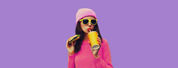 Wall Mural - Portrait of stylish young woman drinking juice eating burger fast food on purple studio background