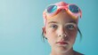 © iuricazac - Young girl with blue eyes wearing pink swim goggles and cap with water droplets on her face against a light blue background.
