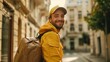 © iuricazac - Smiling man in yellow jacket and cap carrying a brown backpack standing on a city street with buildings in the background.