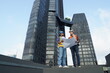 © Supavadee - Foreman engineer and contractor engineer wearing reflective jacket, engineering helmet, holding blueprints, standing on the rooftop of a tall building in the background.