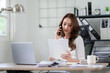 © Wasana - Confident businesswoman discussing work on phone while checking documents in home office, work from home concept