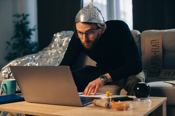 Conspiracy theory. A man in a tinfoil hat looks for signs while watching a video on a laptop.