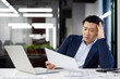 © Liubomir - Confused korean male working with documents in front of portable laptop in office environment. Exhausted businessman dressed in dark suit reading important papers indoors.