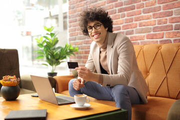 Sticker - Smiling young man sitting in a cafe using a laptop computer and holding a credit card