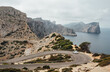 Lonely small white car driving by serpentine curved asphalt mountain road near Lighthouse of Cap de Formentor with beautiful seascape with rocky coast. Mallorca Island, Balearic Islands, Spain.