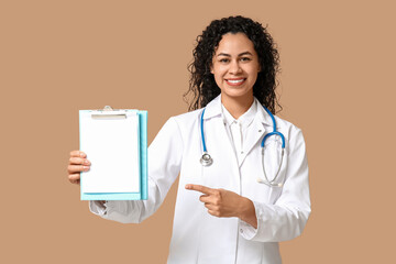 Poster - Beautiful young happy African-American female doctor with stethoscope pointing at clipboard on brown background