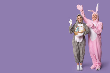 Wall Mural - Beautiful young couple in bunny costume with Easter basket pointing at something on purple background